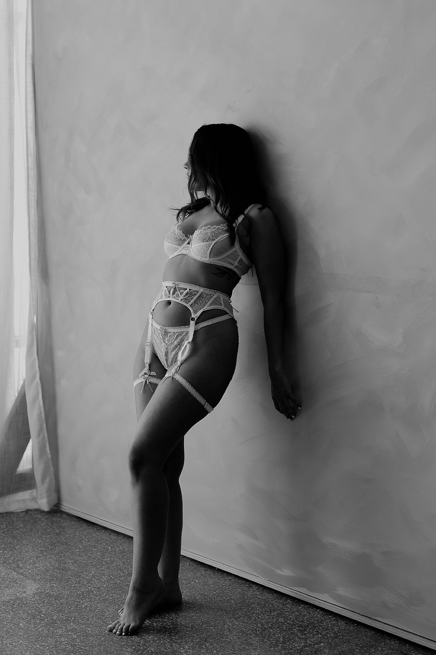 woman leans against studio wall in lingerie after finding the boudoir photographer for you